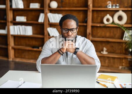 Un homme d'affaires noir concentré a fait des coups de lèvres assis devant un ordinateur portable, mis son menton sur les mains, travaillant sur l'ordinateur avec des documents sur le bureau, pensant aux prochaines étapes, plans, regarde l'écran Banque D'Images