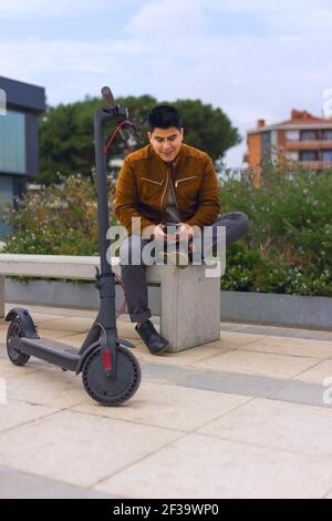 jeune garçon dans une veste marron et un pantalon gris assis sur un banc parlant sur le téléphone portable avec le scooter électrique à l'avant.(mise au point sélective dans scou Banque D'Images