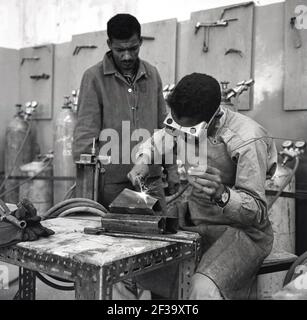 Années 1960, historique, tuteur supervisant un apprenti d'ingénieur masculin portant des lunettes de sécurité, sittnig par une table basse soudant un objet métallique, Arabie Saoudite. La découverte de pétrole dans le pays a vu la croissance rapide de l'industrie pétrolière et le besoin subséquent de travailleurs qualifiés. Banque D'Images