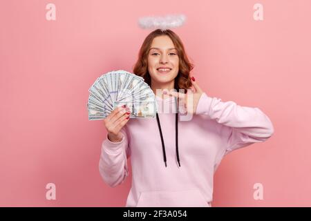 Portrait d'une adolescente enjouée à cheveux bouclés avec halo sur la tête pointant du doigt sur un tas de billets de banque en dollars et souriant, profit. St. Intérieure Banque D'Images