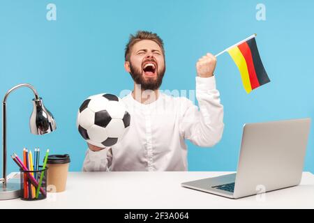 Un fan de football à barbe extrêmement enthousiaste se réjouit sincèrement, soutenant l'équipe favorite tenant le ballon et le drapeau allemand, en regardant le football sur le lieu de travail Banque D'Images