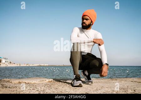 Sportif afro-américain sportif en écouteurs assis pendant l'entraînement à la plage Banque D'Images