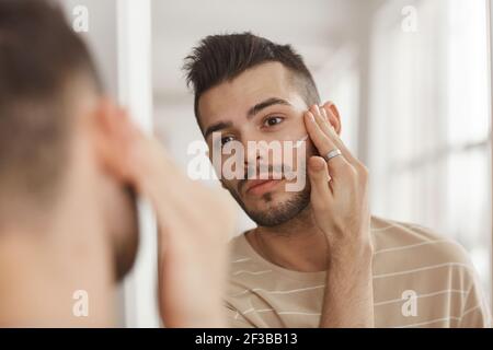 Portrait d'un jeune homme appliquant de la crème pour le visage tout en regardant dans le miroir pendant la routine de soin de la peau du matin, espace de copie Banque D'Images