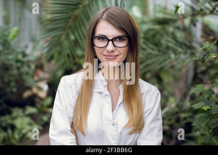 Gros plan de la jeune femme caucasienne, scientifique, botaniste, agronome, portant des lunettes et un manteau blanc de laboratoire, posant à la caméra au-dessus des palmiers Banque D'Images