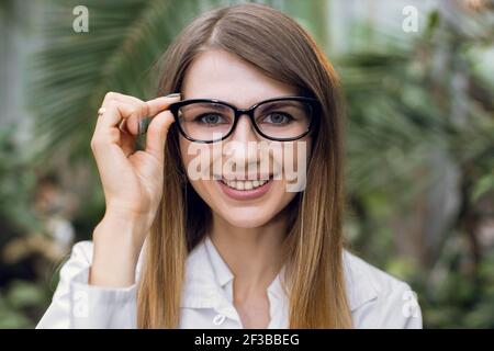 Jolie bonne femme de race blanche agronome, botaniste, portant des lunettes et un manteau de laboratoire blanc, travaillant en serre et regardant la caméra. Gros plan Banque D'Images