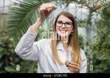 Jeune femme blonde assez souriante scientifique agronome portant un manteau blanc de laboratoire, posant à la caméra en serre avec des plantes tropiques, tenant un tube à essai et Banque D'Images