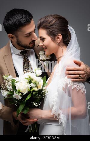 élégant marié arabe embrassant la mariée heureuse tenant le bouquet de mariage isolé en gris Banque D'Images