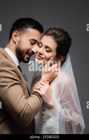 joyeux homme arabe embrassant le cou de jolie mariée souriante isolée sur gris Banque D'Images
