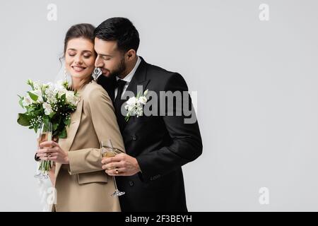 elégant homme arabe avec verre à champagne embrassant les mariés heureux tenue bouquet de mariage isolé sur gris Banque D'Images