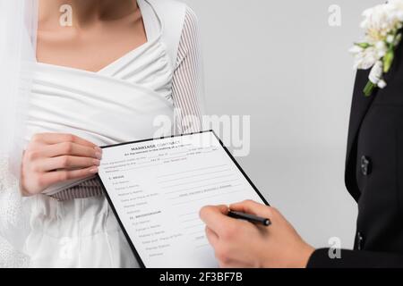 Vue rognée de la mariée tenant le contrat de mariage près de marié avec stylo sur un premier plan flou isolé sur gris Banque D'Images