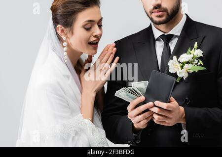 Mariée excitée regardant le marié avec des dollars et portefeuille isolé en gris Banque D'Images