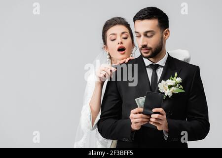Mariée enthousiaste regardant le portefeuille avec l'argent entre les mains de marié musulman isolé sur gris Banque D'Images