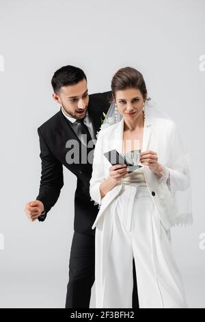 Un groom arabe enthousiaste qui regarde le portefeuille et l'argent entre les mains de mariée isolée sur gris Banque D'Images