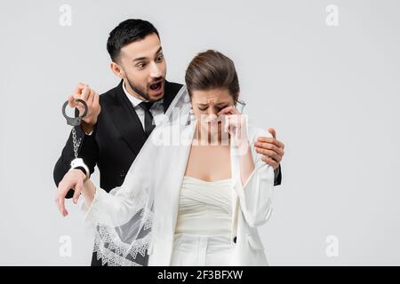 Un marié arabo-arabe enthousiaste embrassant une mariée qui pleure dans des menottes isolées sur gris Banque D'Images