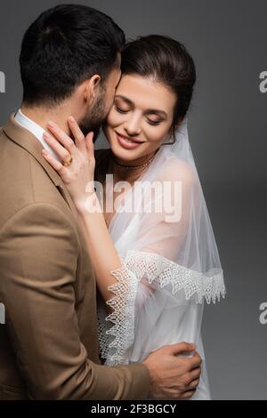mariée souriante aux yeux fermés embrassant le fiancé musulman isolé gris Banque D'Images