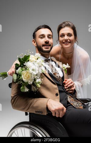 Marié arabe assis en fauteuil roulant près d'une mariée souriante isolée gris Banque D'Images