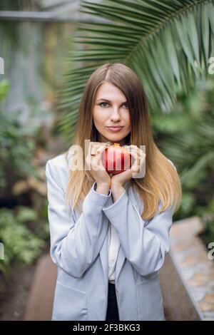 Jeune femme élégante et tendance en veste grise, posant à l'intérieur sur des plantes exotiques arrière-plan dans la serre, tenant la grenade rouge dans les mains et regardant Banque D'Images