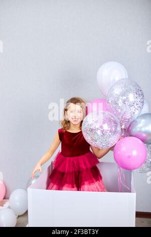 Adorable, joyeuse petite fille en robe rose regarde hors de la grande boîte cadeau avec des ballons à la maison fête streamers, Joyeux anniversaire. Celebrati Banque D'Images