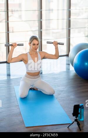 Femme de forme physique mince faisant des exercices avec des haltères tout en étant assis dans avant de l'appareil photo du smartphone Banque D'Images