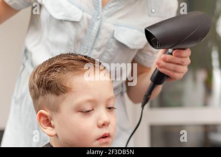 Une mère avec un sèche-cheveux dans sa main dans une robe bleu clair fait les cheveux de son fils à la maison pendant la deuxième période de quarantaine. Foyer sélectif Banque D'Images