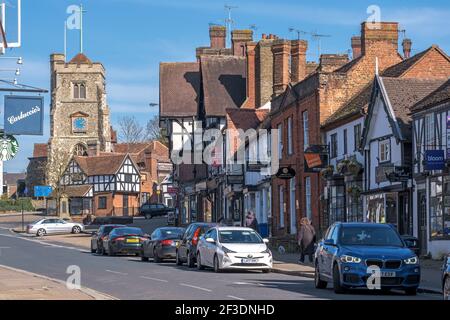 Pinner Village High Street avec l'église médiévale de St Jean-Baptiste au sommet d'une colline, des boutiques, des voitures garées et deux personnes marchant sur le trottoir. Angleterre Banque D'Images