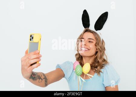 Joyeuses Pâques salutations, jeune femme portant des oreilles de lapin tenant des œufs de couleur souriant rend selfie en utilisant son smartphone debout sur le dos blanc de studio Banque D'Images