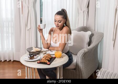 Belle femme tenant une tasse de boisson chaude et regardant le petit-déjeuner est servi sur une table, assis dans un fauteuil de l'hôtel chambre le matin Banque D'Images