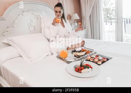 Jolie femme en peignoir tenant un croissant et en sirotant du jus frais du verre tout en étant assis sur un lit moelleux dans la chambre d'hôtel le matin Banque D'Images