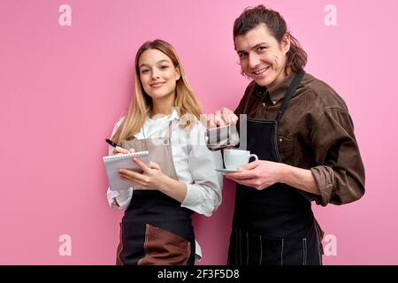 Les jeunes serveurs prêts à servir les clients dans un café ou un restaurant, se posent à la caméra isolée sur fond rose, le serveur mâle versant le café dans le blanc Banque D'Images