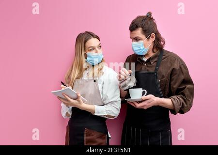 deux jeunes serveurs en tablier et masque discutent des commandes, prêts à servir les clients pendant la quarantaine, serveuse amicale écrivant des notes et parlant Banque D'Images
