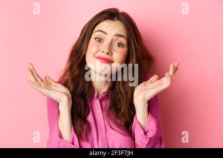 Portrait en gros plan de femme mignonne sans indice, haussant les épaules et souriant avec l'expression du visage inconscient, debout sur fond rose Banque D'Images