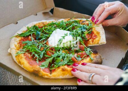 une femme s'assoit dans une voiture et se déchire de la pizza avec fromage burrata et légumes verts avec ongles laqués rouges Banque D'Images