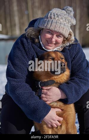 Une femme blonde d'âge moyen, recouverte d'un bonnet tricoté, encadre un mongrel assis sur la neige blanche lors d'une belle journée d'hiver. Banque D'Images