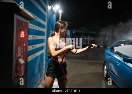 Une jeune femme lave une voiture bleue lors d'un lavage de voiture. Fille posant sur l'appareil photo Banque D'Images