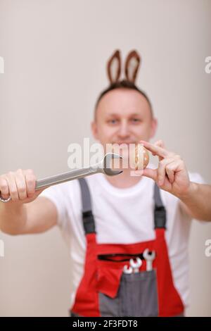 Vacances de Pâques, concept de célébration. Travailleur ou mécanicien souriant drôle porte des oreilles de lapin sur la tête, tenant des clés et peint blanc d'oeuf Banque D'Images