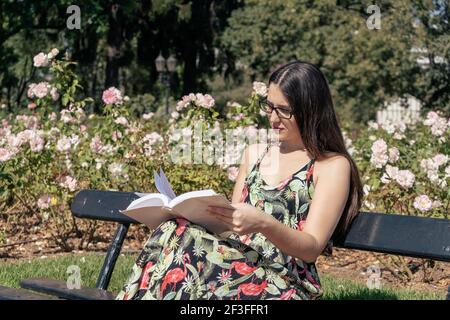 Belle jeune femme latine avec des verres noirs et une robe fleurie dans un parc concentré de lecture d'un livre. Concept de culture. Banque D'Images