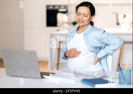 Femme adulte enceinte épuisée assise à son bureau tenant dans le bas du dos et de l'estomac, souffrant de douleurs dans le bas du dos, fatiguée du travail sédentaire avec un ordinateur portable, a besoin d'une pause et de repos Banque D'Images