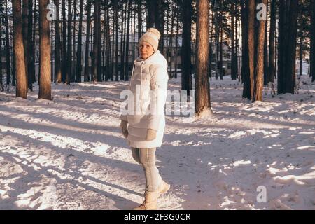 Une femme heureuse d'âge moyen dans une veste chaude, un chapeau et des moufles marche dans un parc hivernal enneigé au coucher du soleil. Repos d'hiver et promenades dans le parc de la ville. Banque D'Images