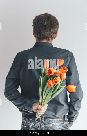 Beau homme avec un bouquet de tulipes orange derrière son dos. Surprise pour votre femme bien-aimée. Concept de vacances romantiques. Saint-Valentin, Fête des mères Banque D'Images