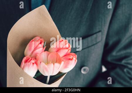 Homme élégant tenant un bouquet de tulipes roses enveloppées de papier artisanal. Surprise pour votre femme bien-aimée. Concept escapade romantique. Mise au point sélective. Banque D'Images