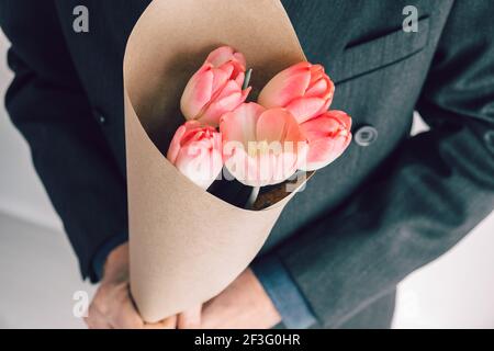 Homme élégant tenant un bouquet de tulipes roses enveloppées de papier artisanal. Surprise pour votre femme bien-aimée. Concept escapade romantique. Mise au point sélective. Banque D'Images