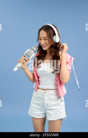 Jeune femme asiatique écoutant de la musique avec un casque, chantant une chanson dans le microphone sur fond bleu. Banque D'Images