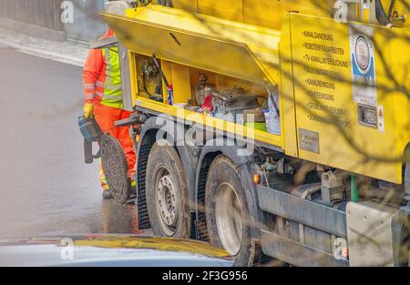 Entretien dans un trou d'homme avec un homme méconnaissable, travaillant derrière le véhicule de nettoyage d'égout sur la rue ouverte. Banque D'Images