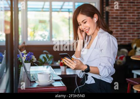 Bonne femme asiatique qui travaille sur un ordinateur portable et écoute de la musique sur votre smartphone avec un casque installé dans le café. Banque D'Images