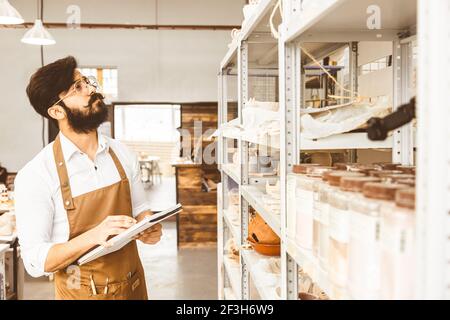 Jeune homme d'affaires séduisant un potter avec une barbe et une moustache travaille dans son atelier. Conserve les enregistrements et transcrites dans un ordinateur portable en inspectant Banque D'Images