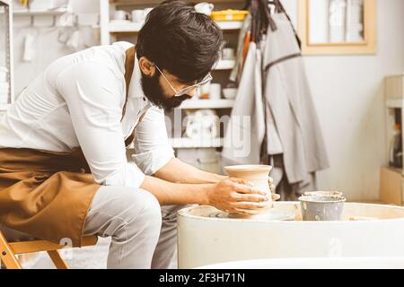 Un jeune potier mâle est engagé dans l'artisanat dans son atelier sur la roue d'un potier et fait un produit d'argile Banque D'Images