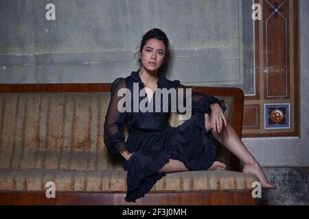 Une belle et solitaire femme avec les cheveux attachés est assise sur un canapé en ruines sans chaussures et portant une longue robe noire, regarder sur l'appareil photo. Banque D'Images
