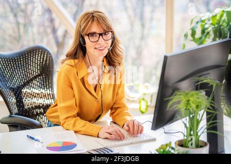 Bonne femme d'affaires mature dans un micro-casque parlant lors d'une conférence téléphonique tout en regardant la caméra. Bureau à domicile. Banque D'Images