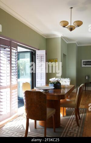 Salle à manger avec table et chaises en ronce art déco. Lumière filtrée à travers des volets partiellement ouverts jusqu'aux portes-fenêtres | AUCUN | Banque D'Images