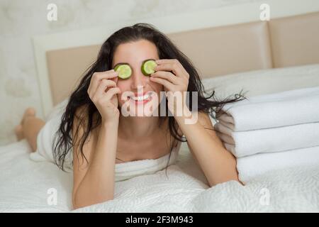 Belle femme souriant au lit. Jeune femme se reposant dans la chambre à coucher avec un masque de tranches de concombre. Portrait de femme à la maison. Soins de la peau le matin. Banque D'Images